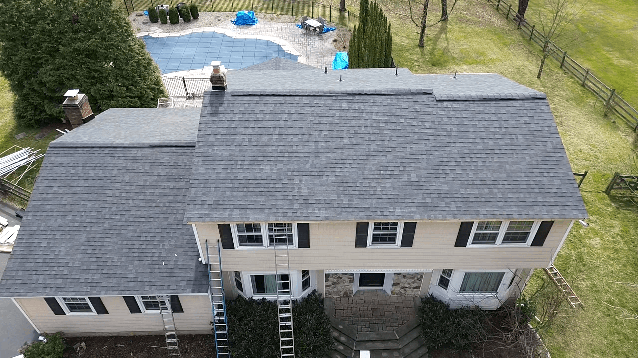 Aerial view of a two-story house with newly installed gray shingle roof, stone patio, and in-ground swimming pool with cover in the backyard