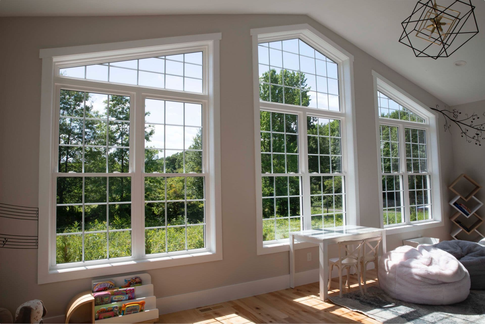 Bright room with large, arched windows showing a green landscape. Inside, soft seating, a small white table, and a rack with children's books create a cozy, inviting space.