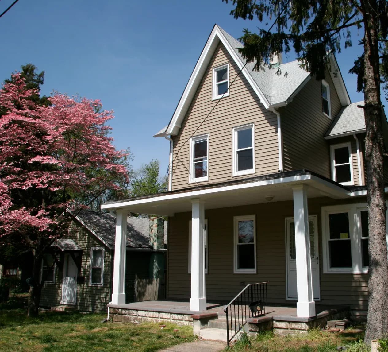 A two-story brown house with a gabled roof and white porch stands beside a blossoming pink tree under a clear blue sky, conveying a serene, springtime feel.