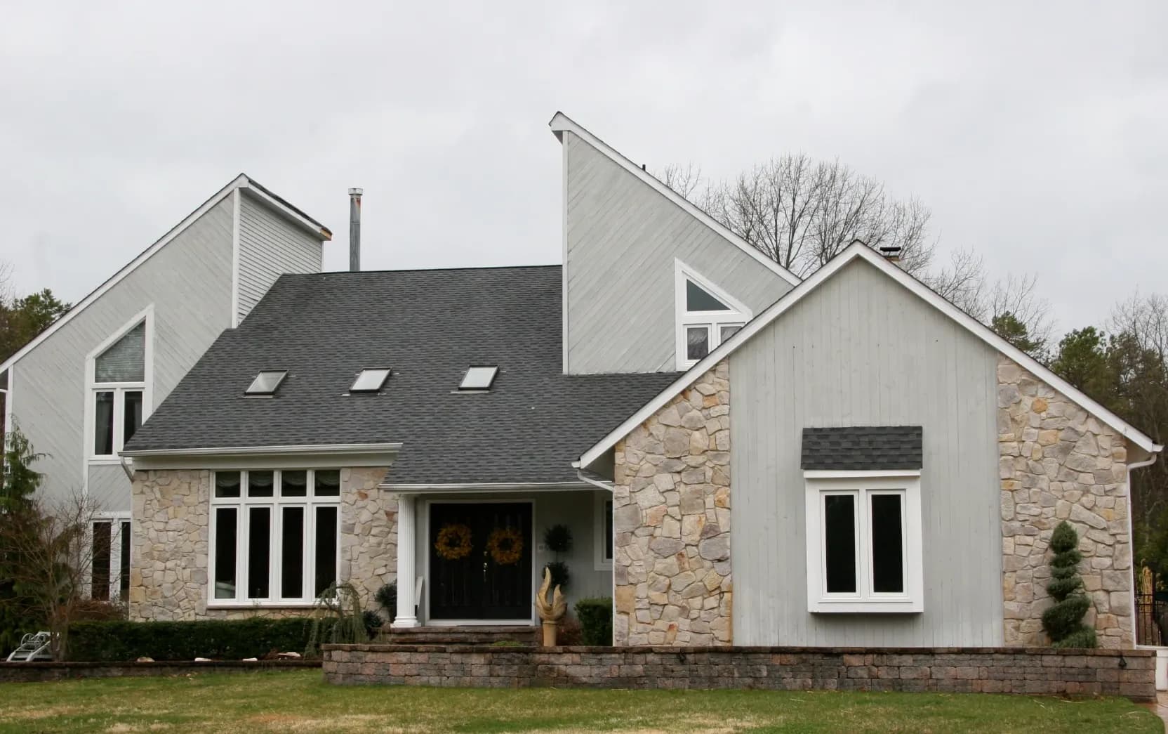 A modern house with stone and wood siding, large windows, and a dark gray roof stands in a yard. Overcast sky adds a calm, serene ambiance.