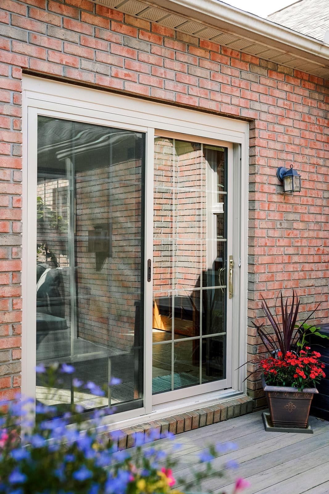 Sliding glass door set in a red brick wall, reflecting a cozy indoor scene. Vibrant potted flowers on a wooden deck add a lively touch.