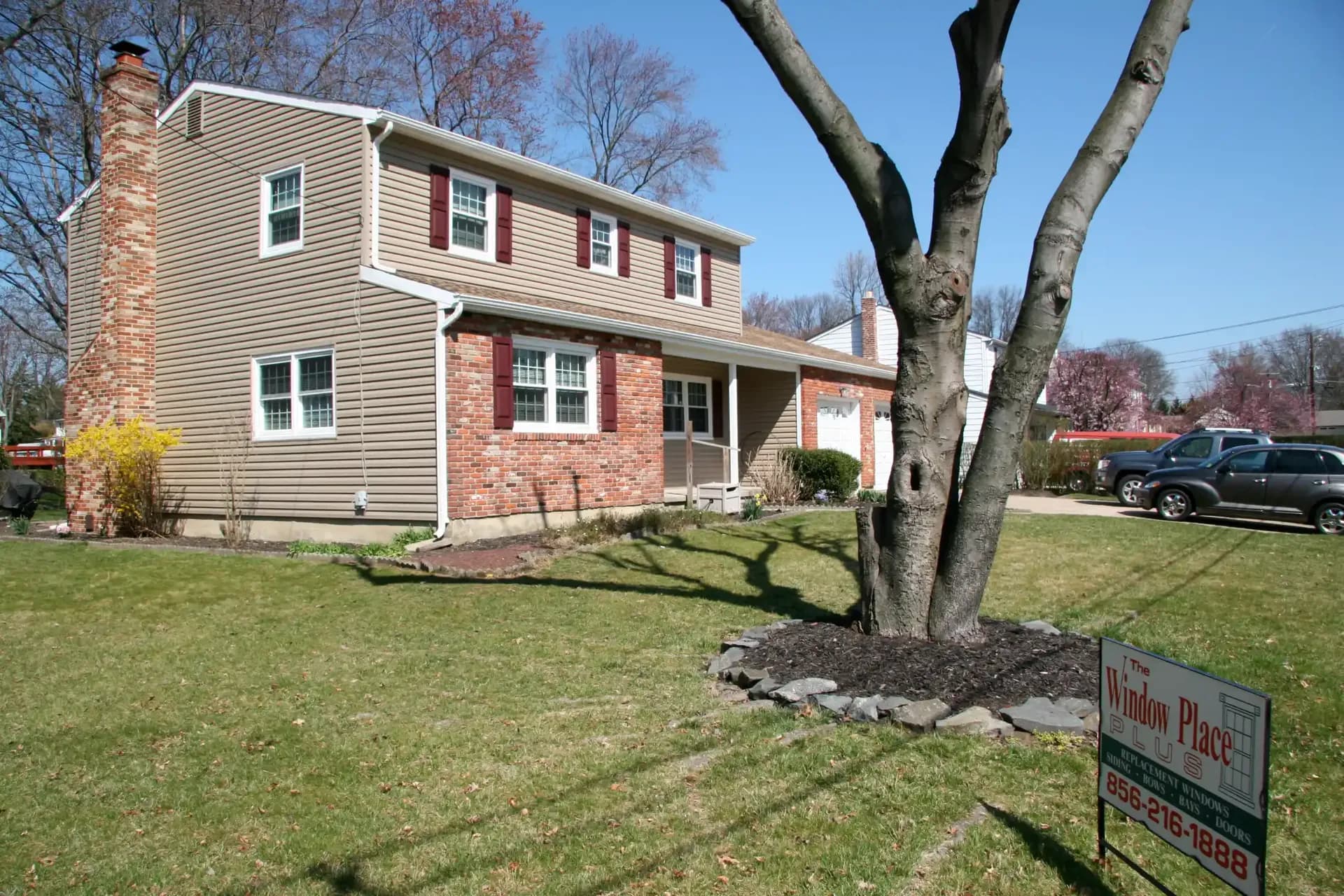 Two-story brick house with beige siding, surrounded by a neat lawn and leafless trees. A sign for a window company is in the foreground.