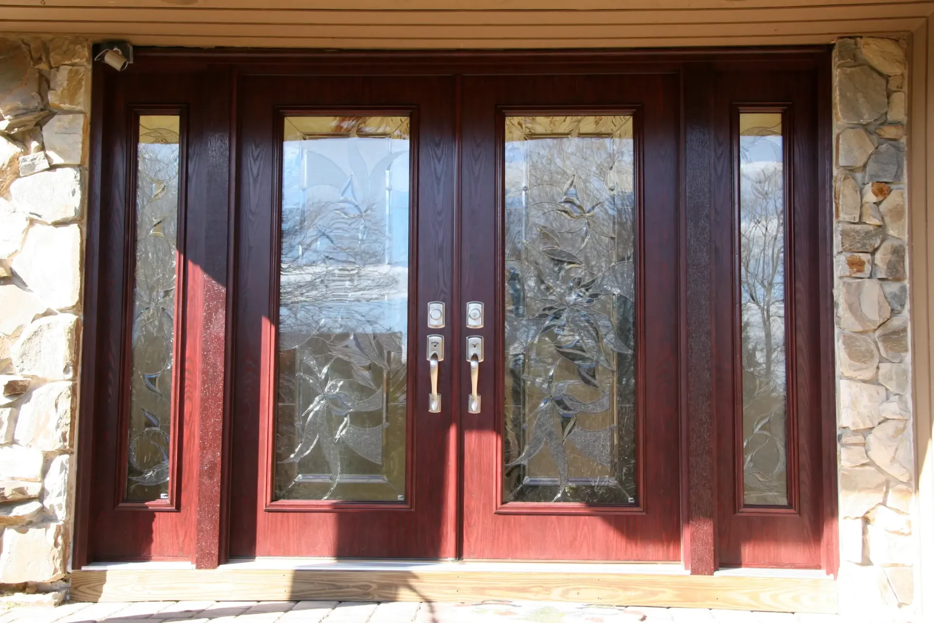 Red double front doors with intricate frosted glass panels, flanked by stone walls. Sunlight creates a warm and inviting atmosphere.