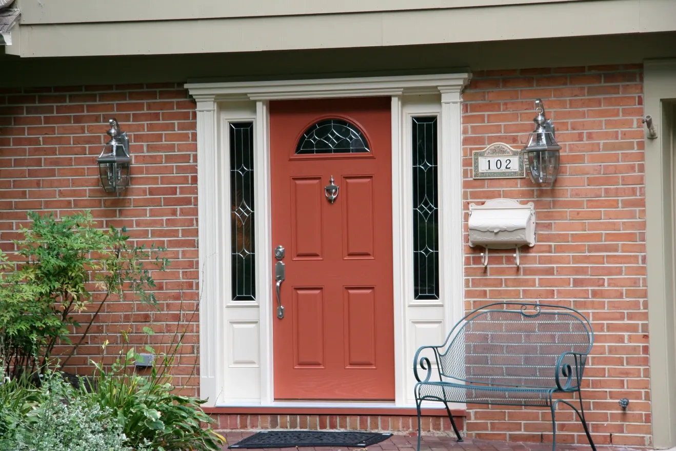 Alt text: "Red front door with white trim set in a brick wall, flanked by two decorative glass panels. A silver mailbox and house number 102 are on the right, with a metal bench below. Lush green plants are on the left."