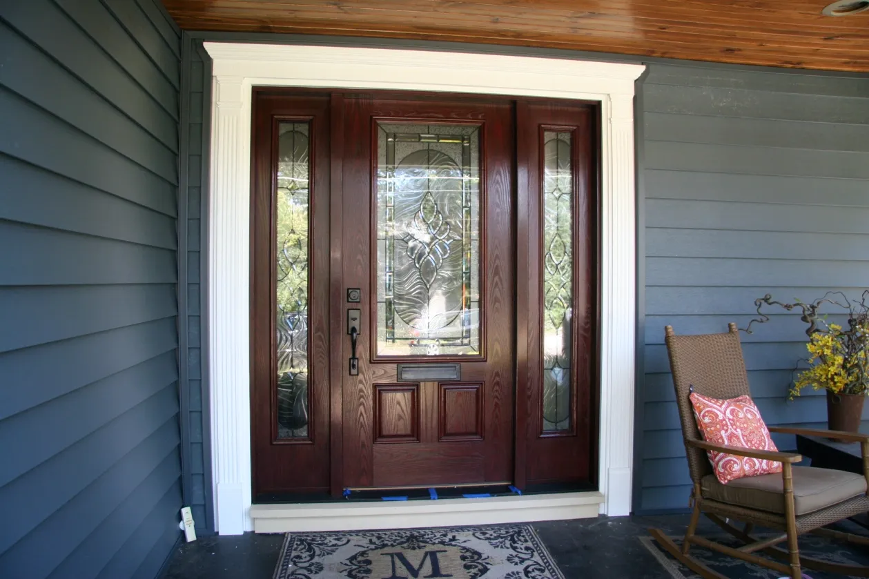 Elegant wooden door with decorative glass panes set in a blue-paneled exterior. Right side features a wicker chair and yellow flowers, evoking a cozy ambiance.