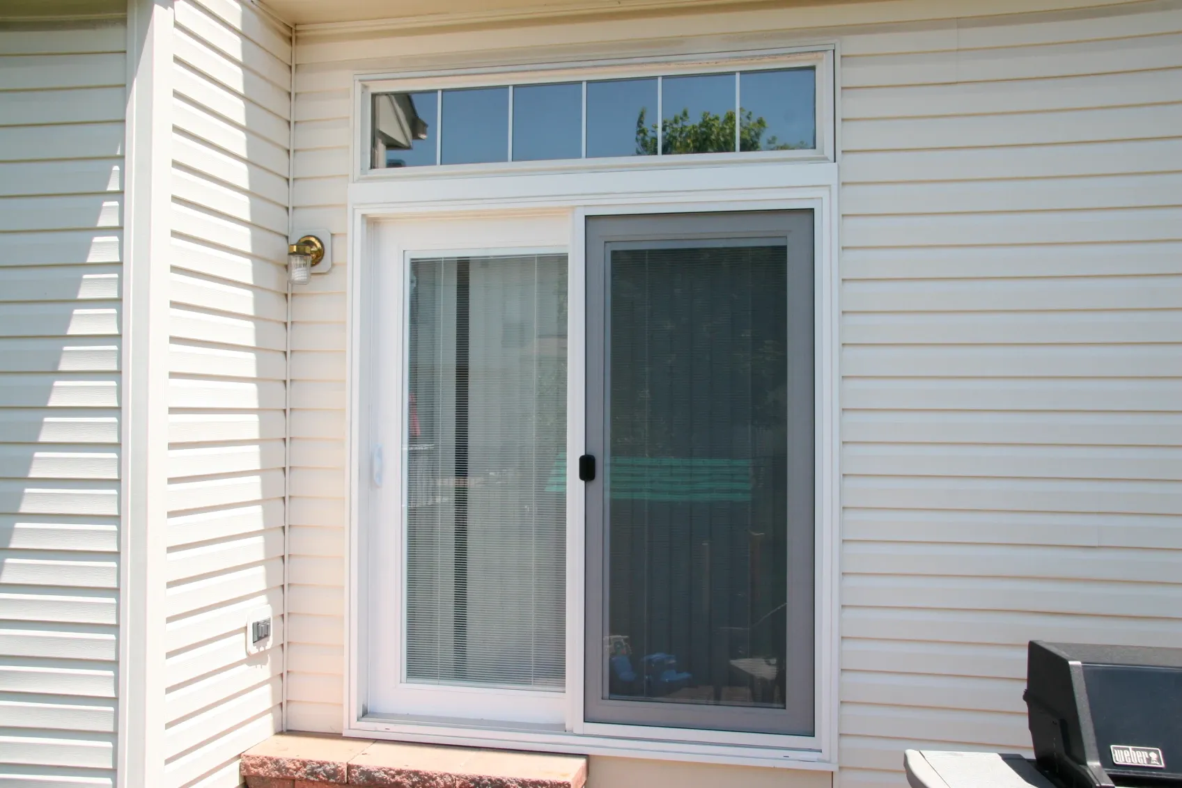 Sliding glass patio door with vertical blinds partially open, set in white siding with a transom window above. A small outdoor grill is nearby.