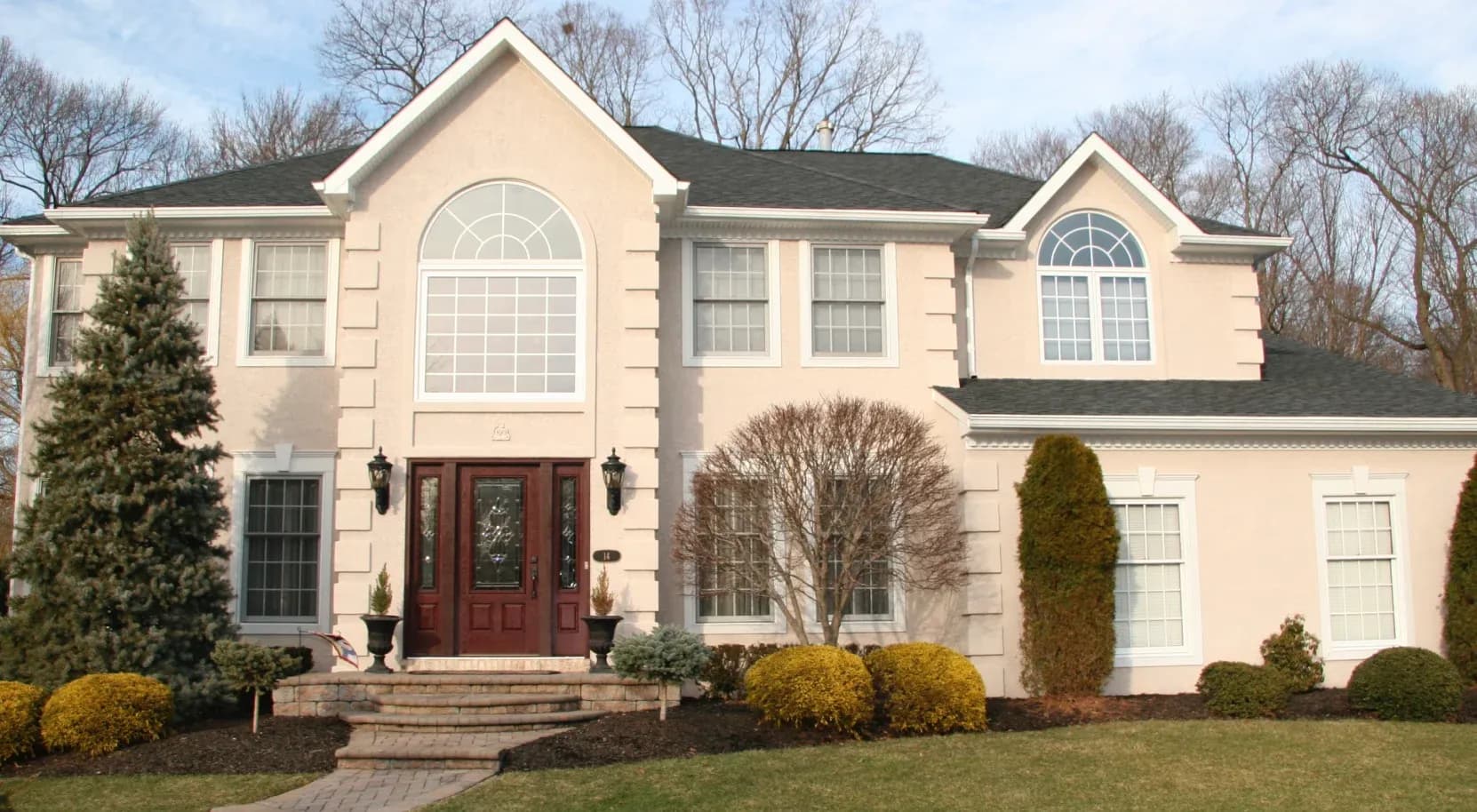 Elegant two-story beige house with large windows and decorative shrubs. Brick steps lead to a red front door. A serene suburban setting.