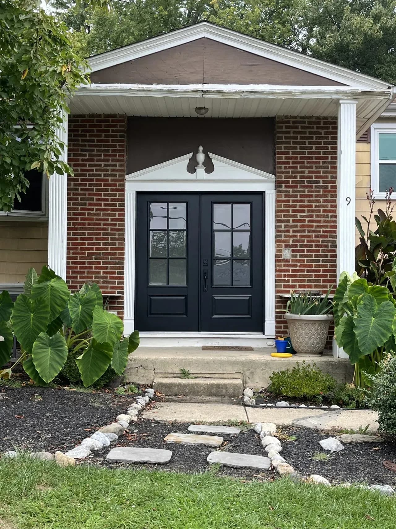 A brick house entrance with black double doors, flanked by white columns. Large green plants and a stone path lead up to the front steps.