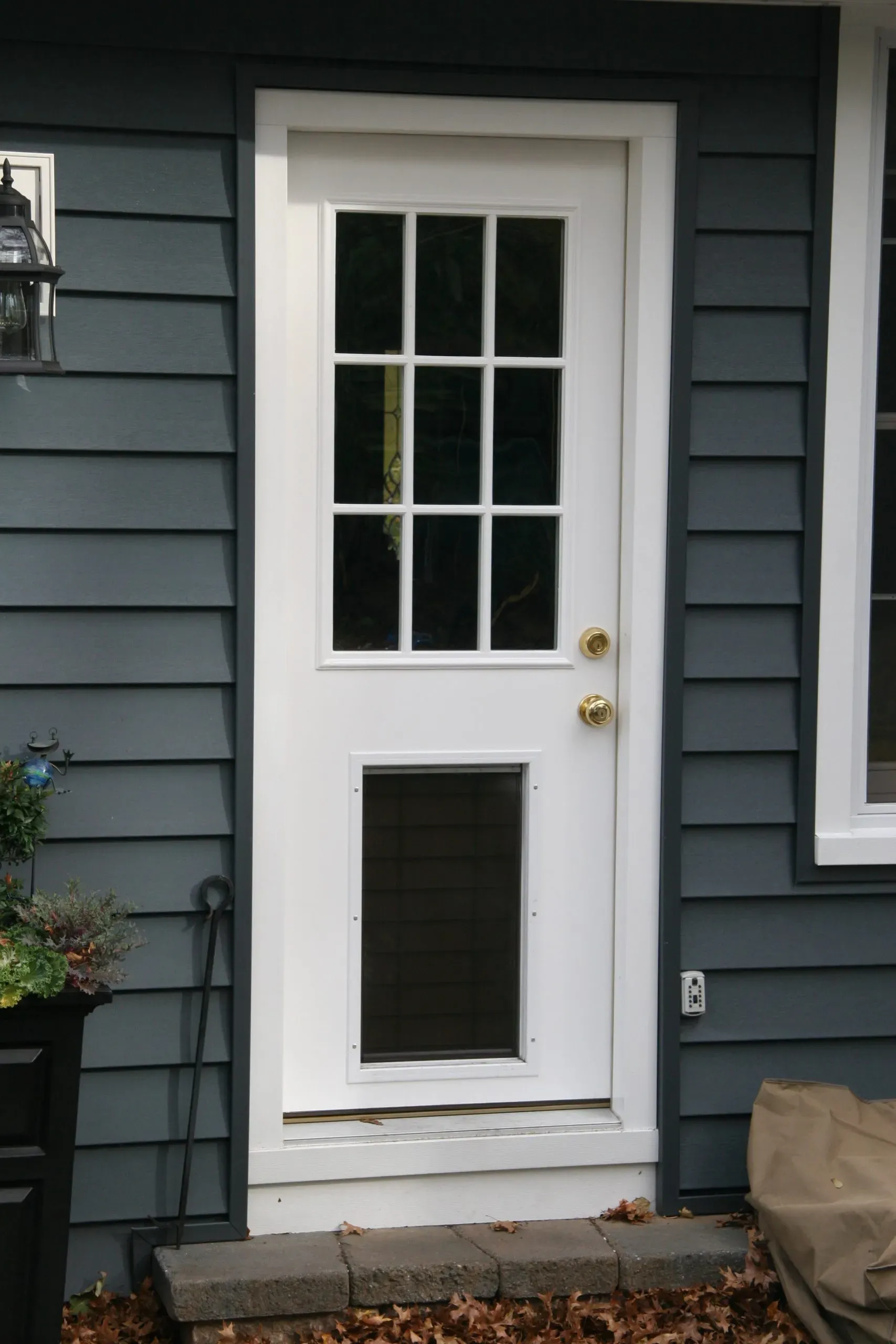 A white exterior door with glass panels and a dog door at the bottom, set against gray siding. A lantern and a potted plant add a welcoming touch.