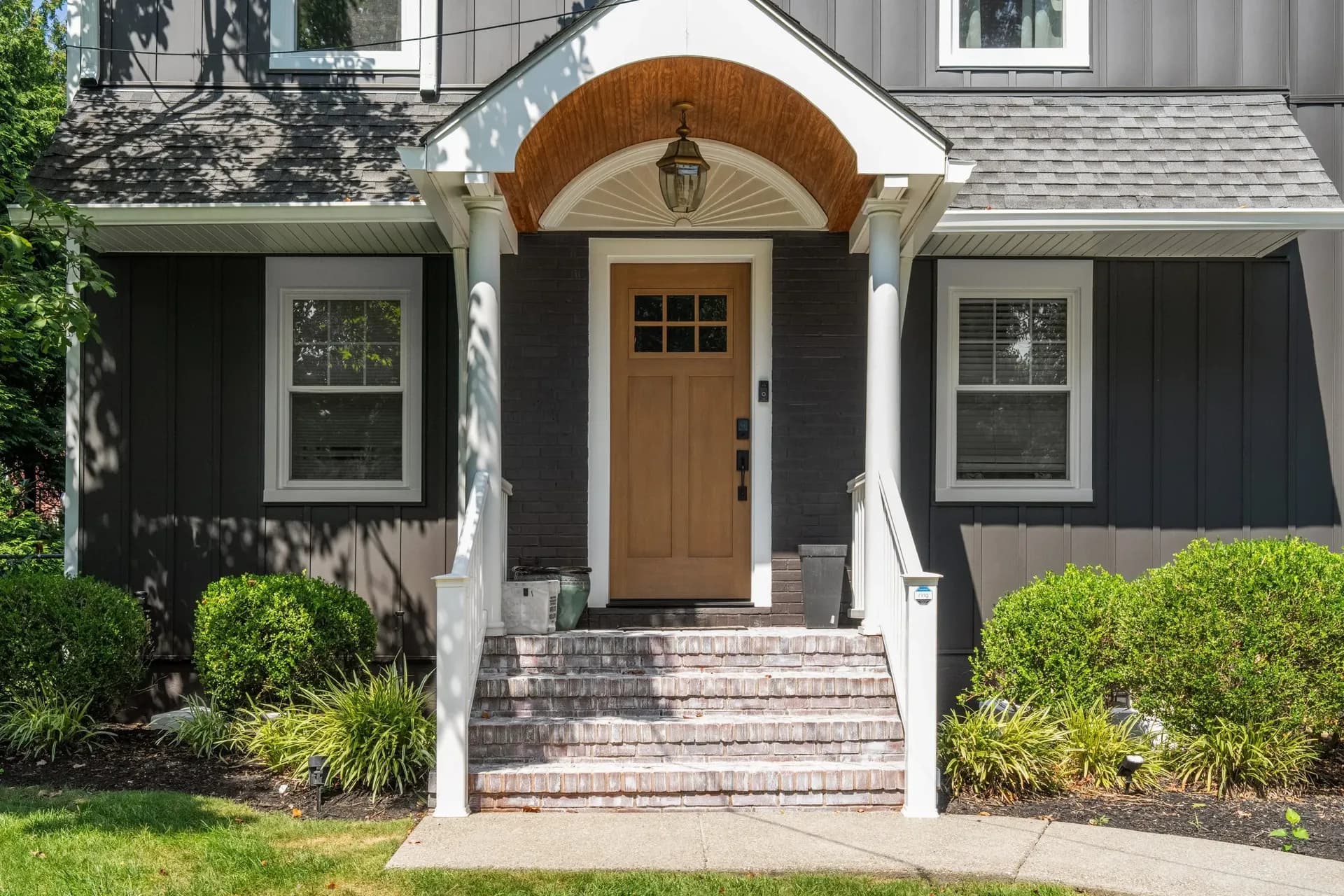 A front porch with a wooden door, white columns, and steps, surrounded by shrubs and grass.