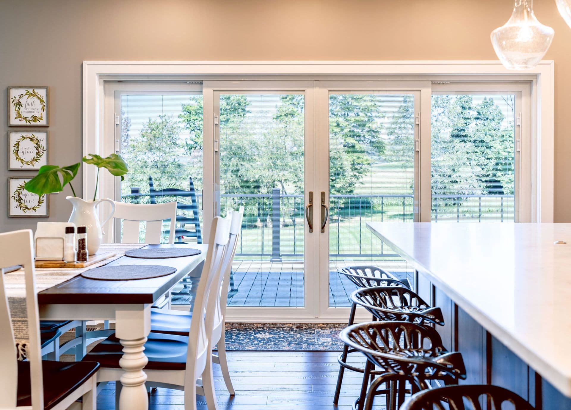Bright kitchen with a large island and white chairs facing glass patio doors. Outdoor greenery is visible, creating an inviting and open atmosphere.