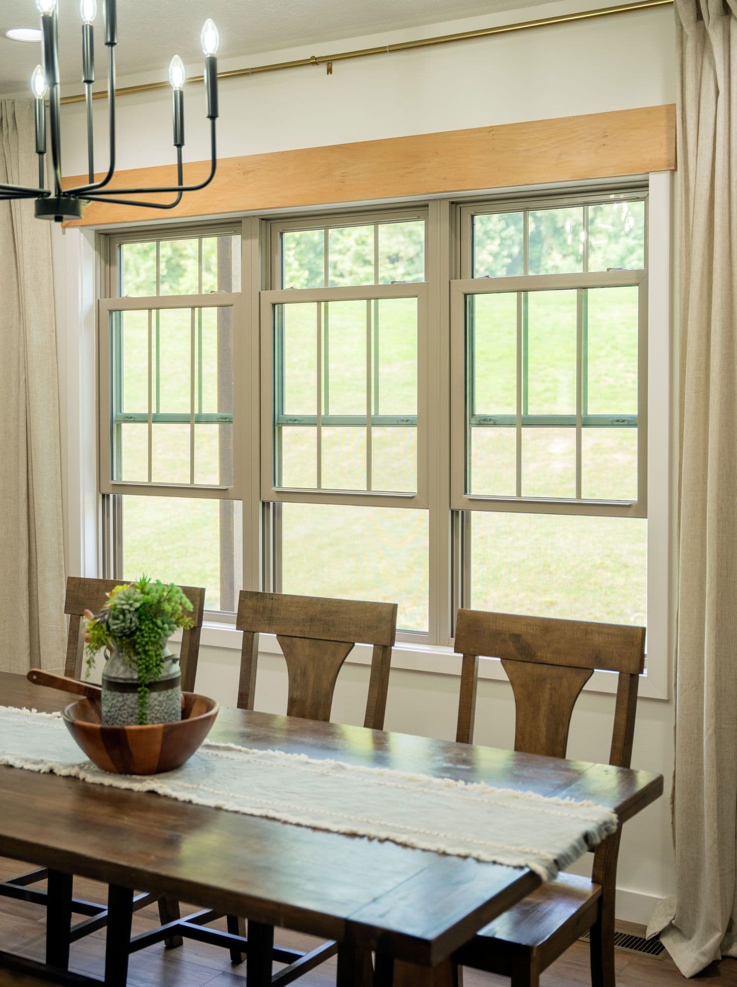Dining room with large windows, wooden table and chairs, a plant centerpiece, white runner, and modern chandelier. Bright, airy, and inviting atmosphere.