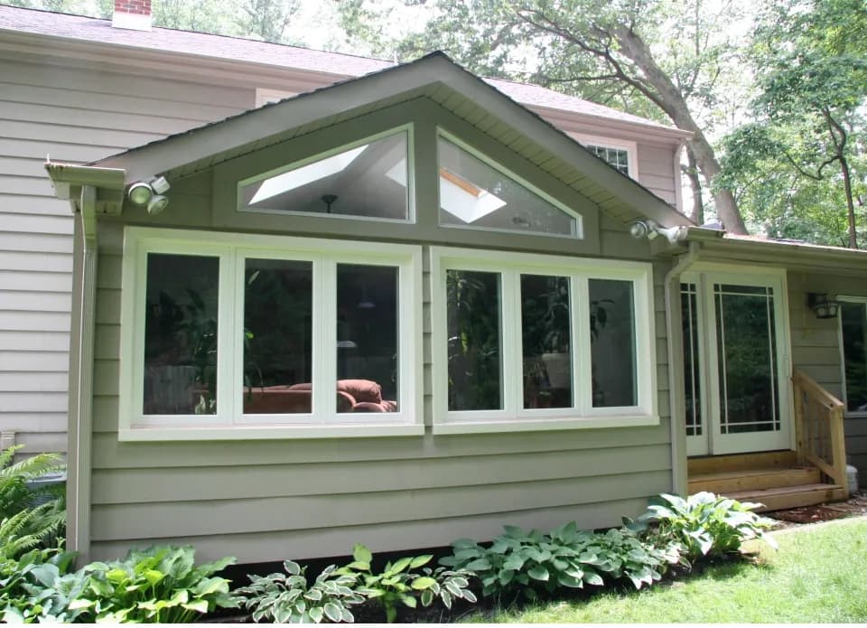 A gray house with a sunroom addition featuring large windows and a triangular top window. Lush greenery surrounds the base, creating a serene atmosphere.