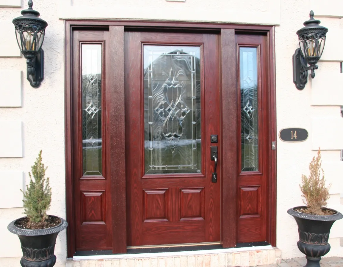 A wooden front door with intricate glass panels, flanked by ornate black lanterns and two potted plants, creates an elegant, welcoming entryway.