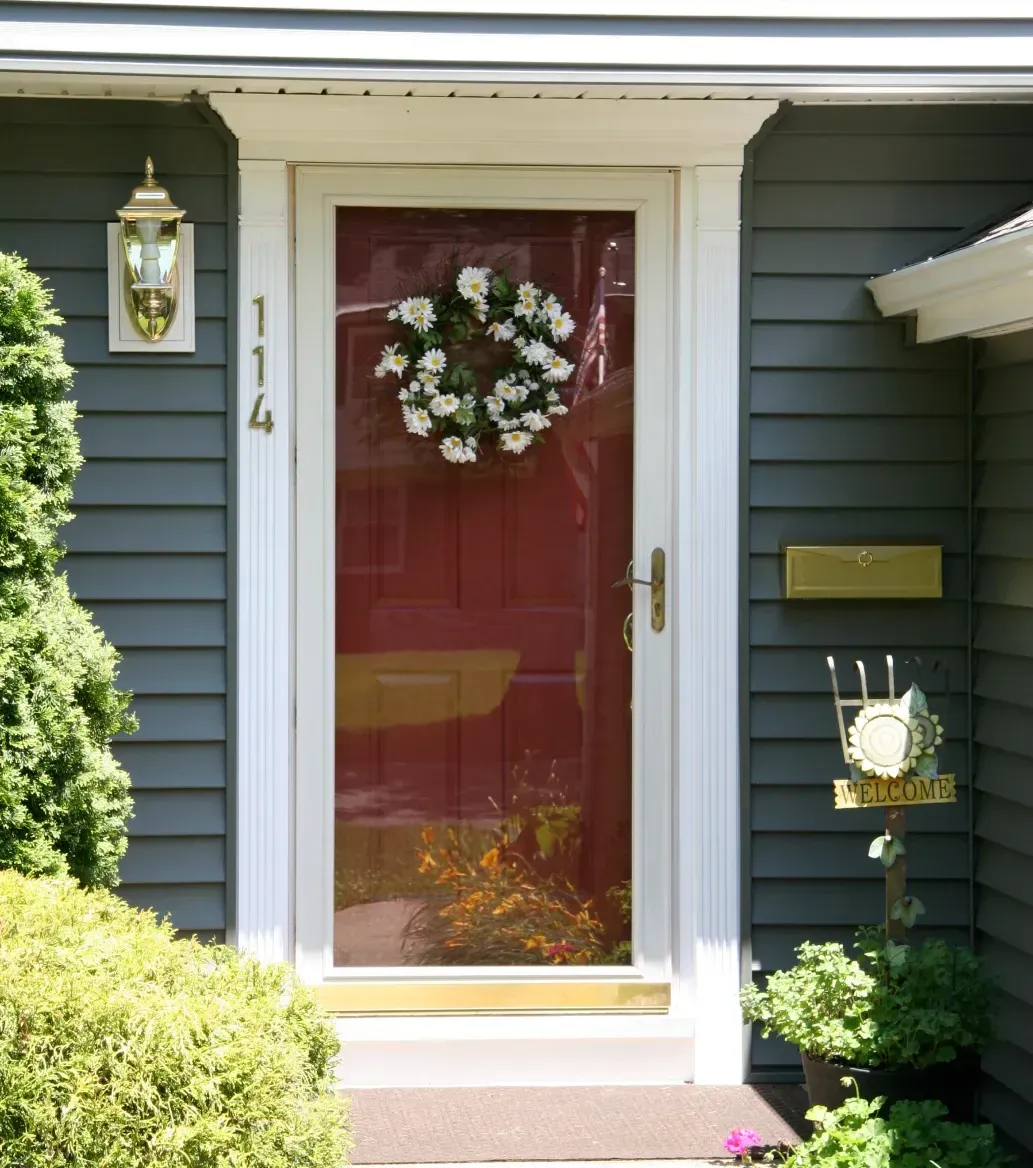 Front door with a red tint, adorned with a white daisy wreath. Gray siding surrounds the door, with a gold mailbox and a "Welcome" sign nearby.