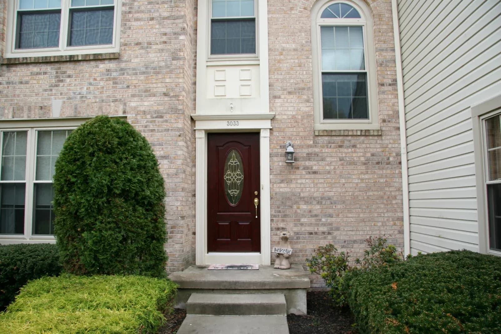 Brick house facade with a dark red front door featuring decorative glass. Flanked by tall windows, a manicured bush, and "Welcome" statue enhance warmth.
