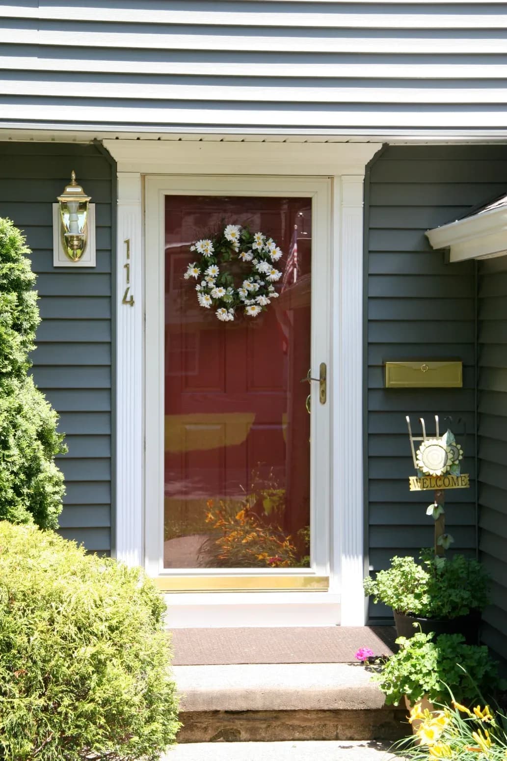 A welcoming front door with a glass storm door adorned by a white floral wreath. Blue-gray siding, potted plants, and a "Welcome" sign enhance the inviting scene.