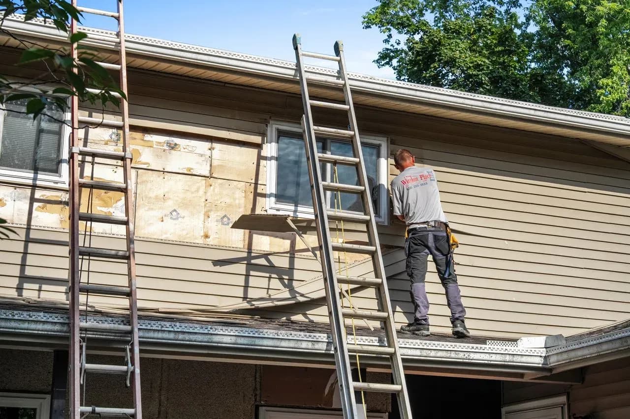 A worker stands on a ladder repairing siding near a second-story window of a house. Two ladders are positioned against the structure, and the scene is outdoors with trees visible.