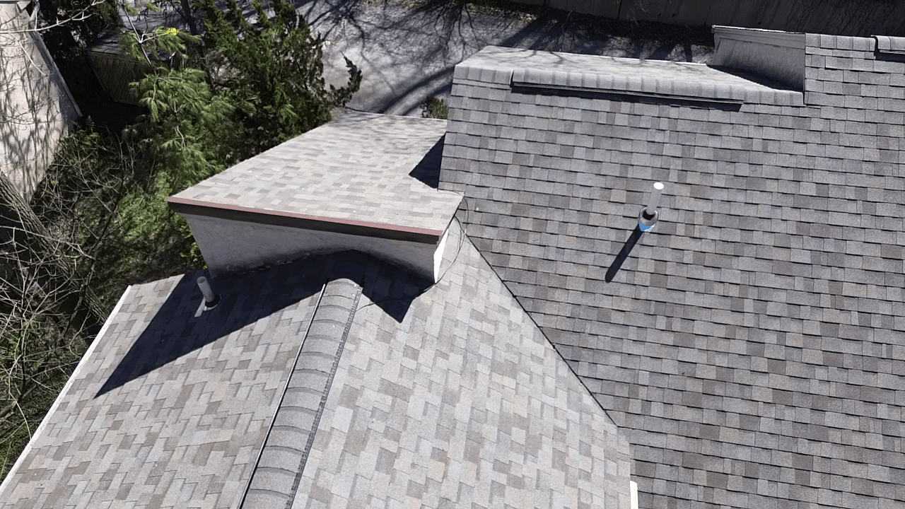 Aerial view of residential roofing showing asphalt shingles, gutters, a skylight, and an adjacent flat-roofed structure with surrounding trees and shadows
