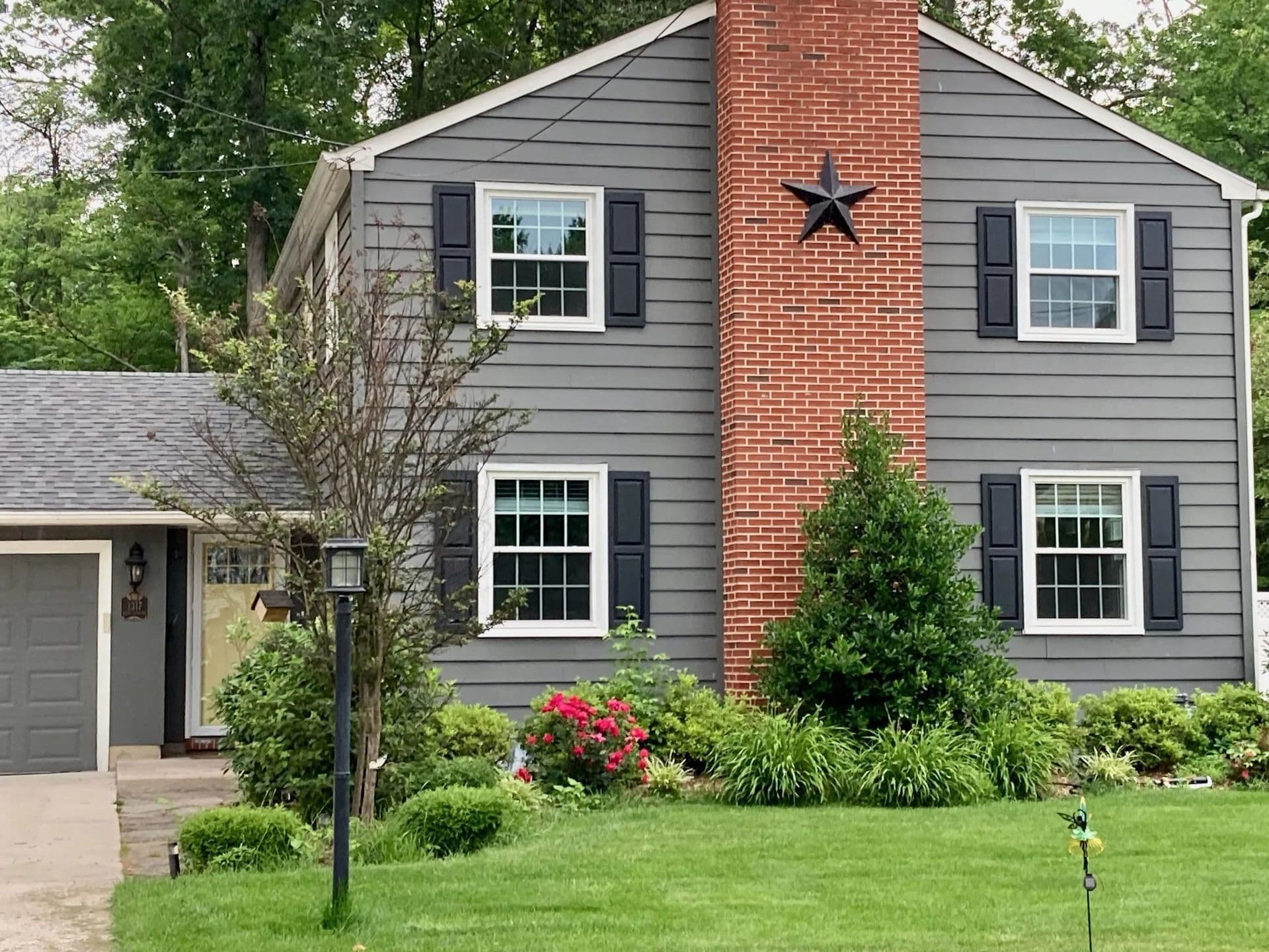 Gray two-story house with black shutters, a red brick chimney, and a star decoration. Lush greenery, colorful flowers, and a neat lawn enhance the cozy, welcoming vibe.