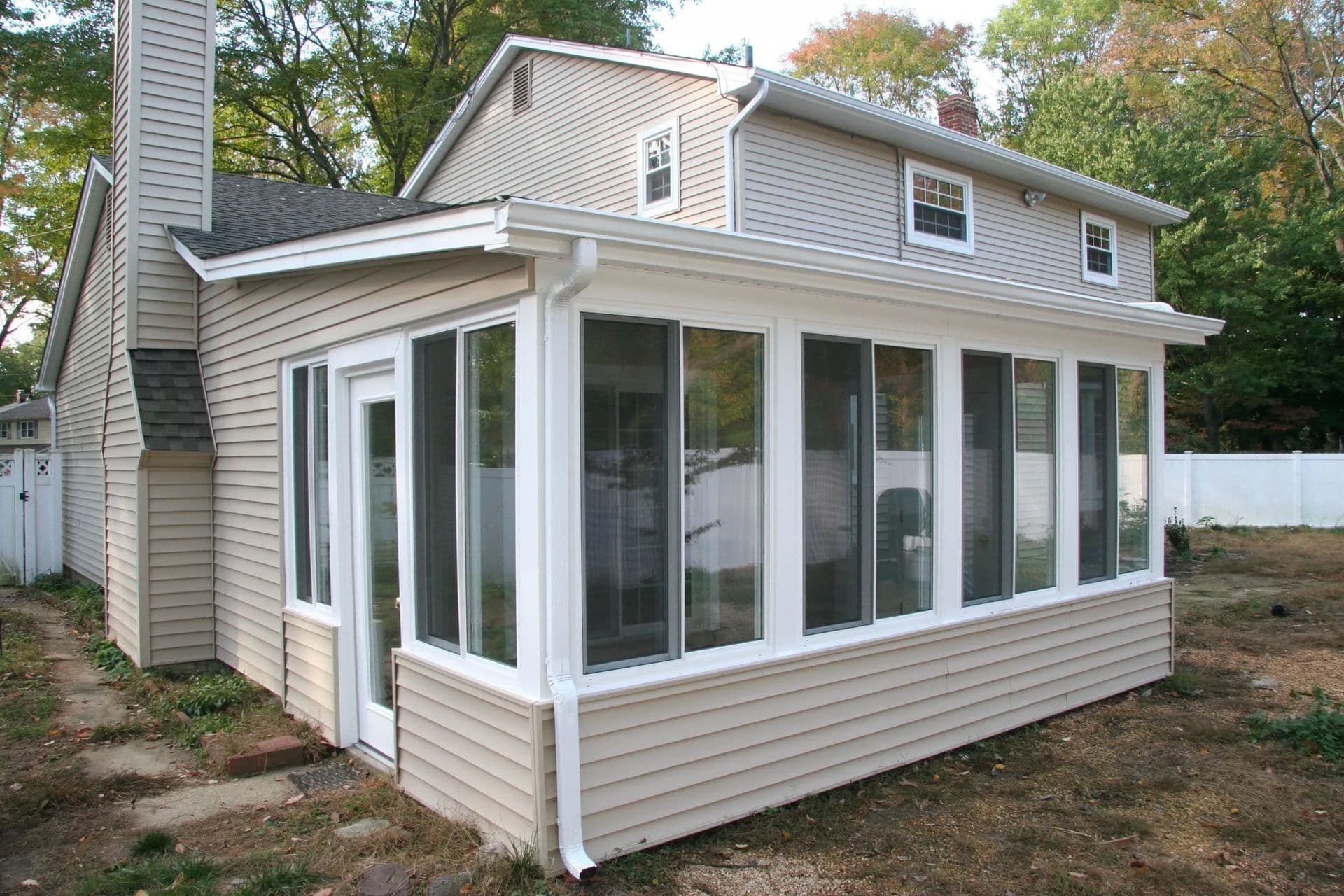 Two-story beige house with a sunroom. Large windows, white trim, and a sloped roof. Surrounded by greenery and enclosed by a white fence.