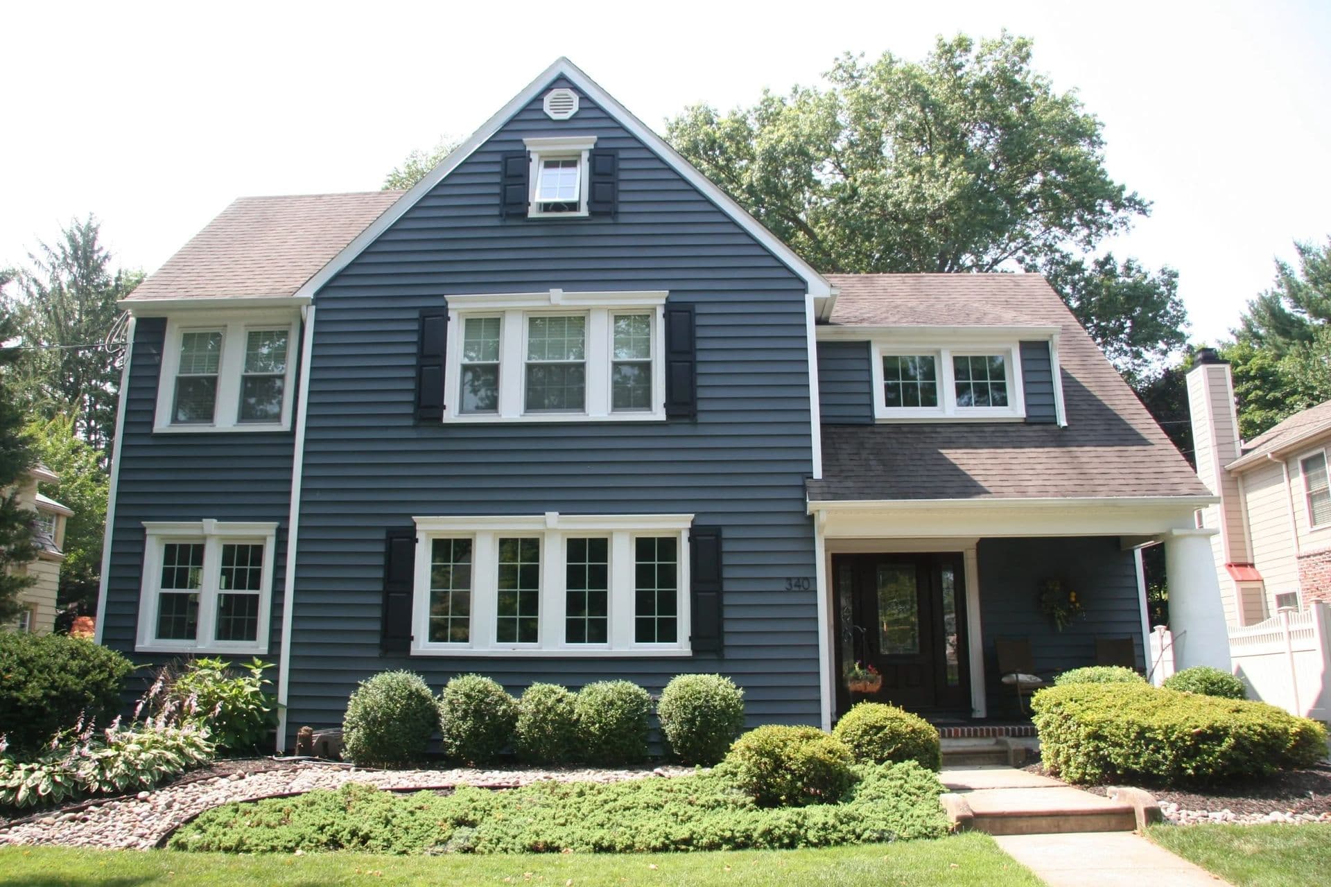 A two-story blue house with white-trimmed windows and black shutters, surrounded by lush green landscaping. The tone is peaceful and welcoming.