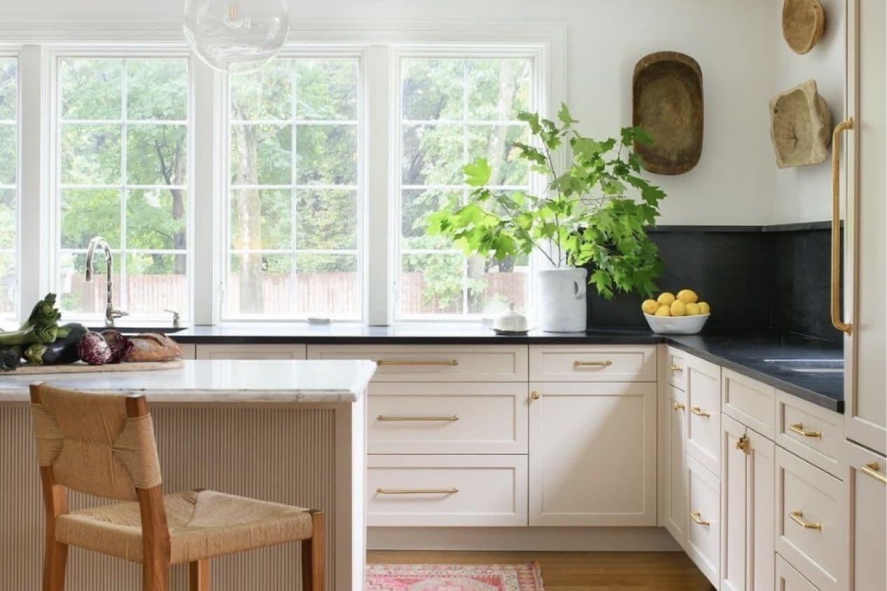 Bright kitchen with large windows and green views. Features cream cabinets, black countertop, leafy plant, bowl of lemons, and woven chair, creating a fresh, airy feel.