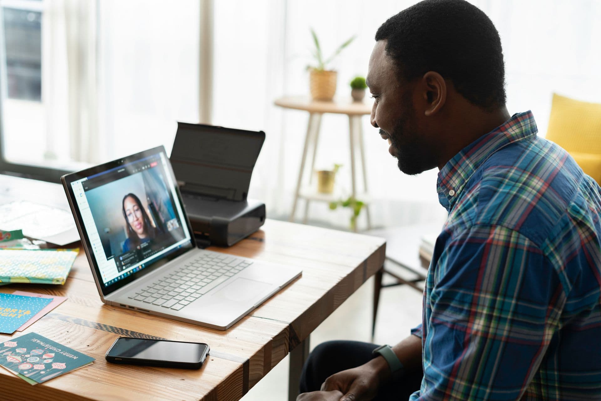 Man in plaid shirt having a video call with a woman on laptop at a wooden desk in a bright home office