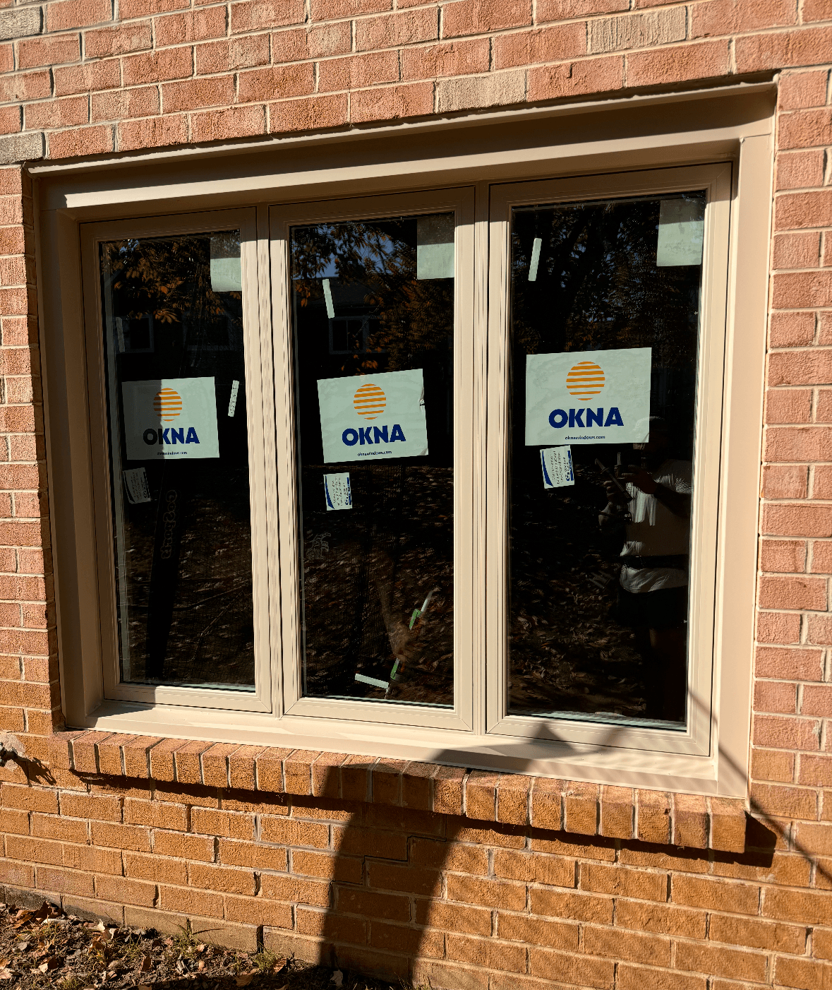 Three-panel window with beige frames installed in a brick wall. Each panel features a label with the "OKNA" logo. Shadows and reflections are visible.