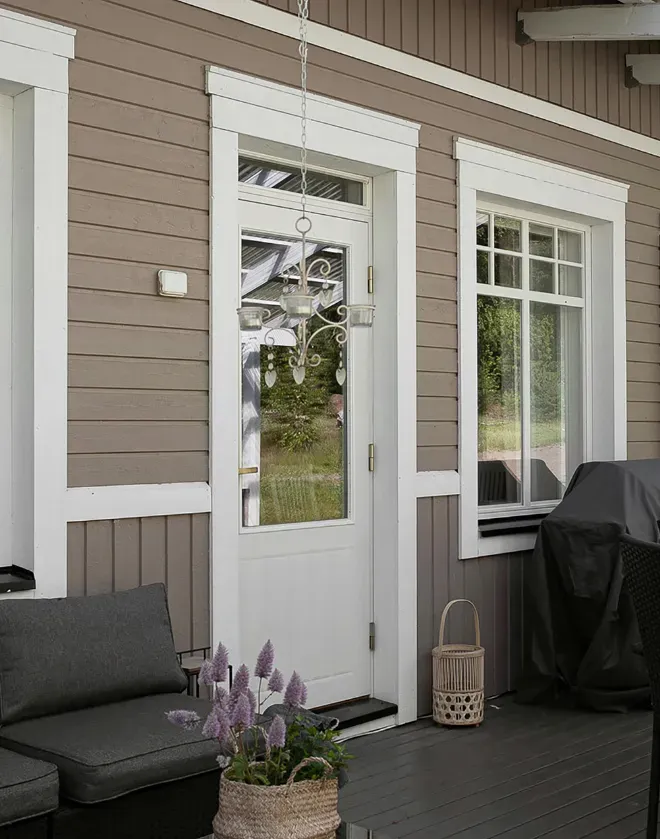 Cozy porch with beige siding, featuring a white door and window. A wicker basket with purple flowers, gray sofa, and elegant hanging light create a welcoming feel.