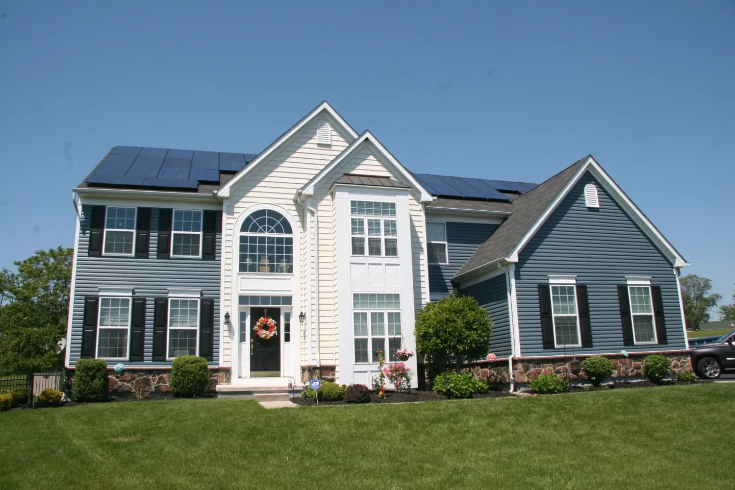 Two-story house with blue and white siding, solar panels on the roof, and a curved entrance. Well-maintained lawn and clear blue sky create a calm ambiance.