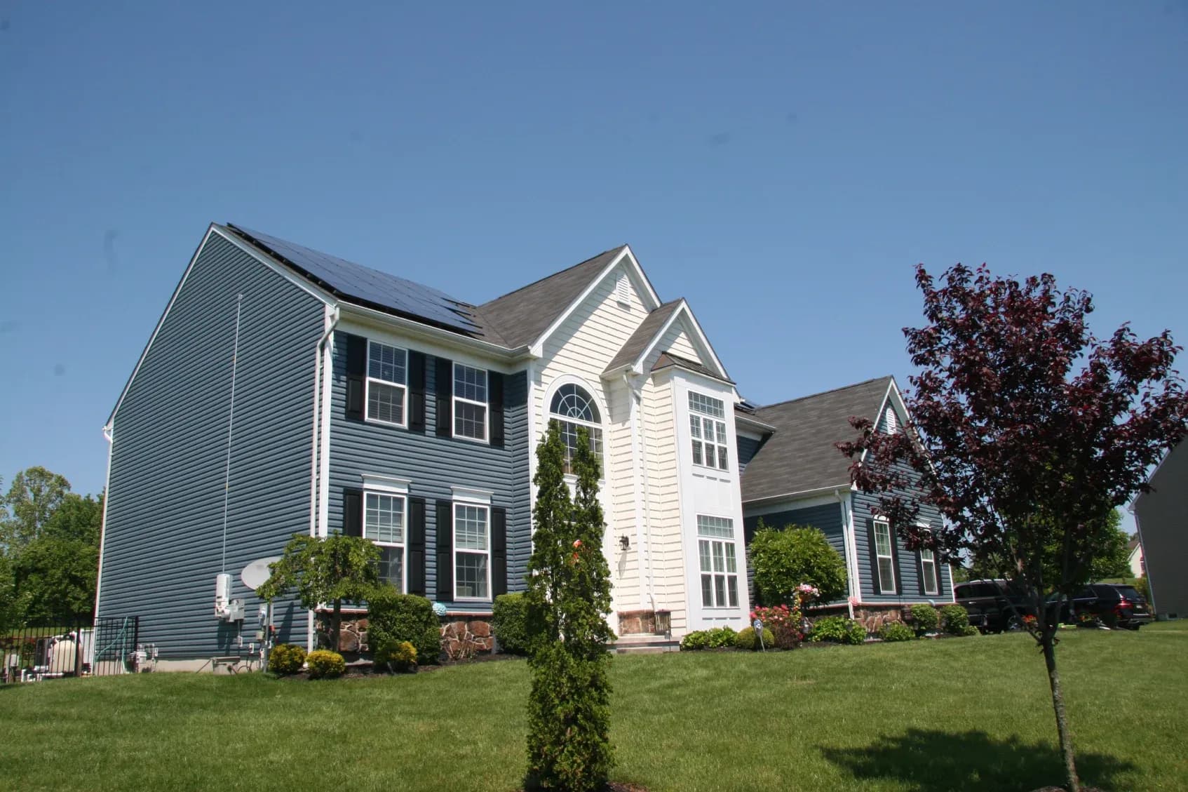 Two-story blue and white house with solar panels on the roof, surrounded by a lush green lawn and trees, under a clear blue sky.