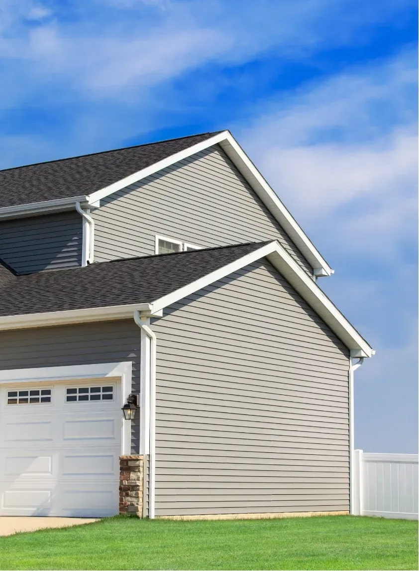 A modern suburban gray house with white trim and a black roof sits under a clear blue sky. The garage door is closed, and a green lawn surrounds it.