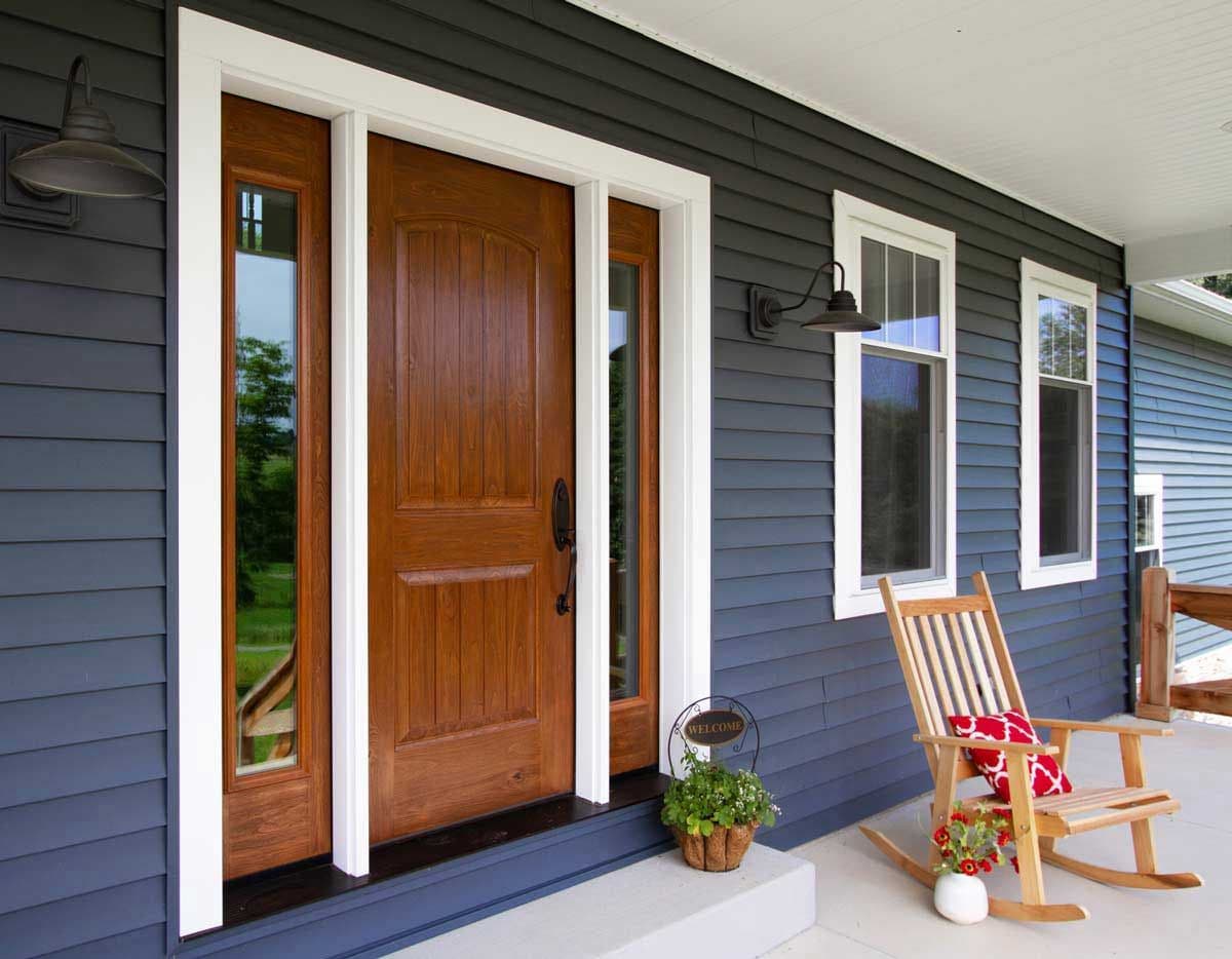 Cozy porch with a wooden front door framed by narrow windows, dark blue siding, and white trim. A rocking chair with red cushions sits beside potted flowers.