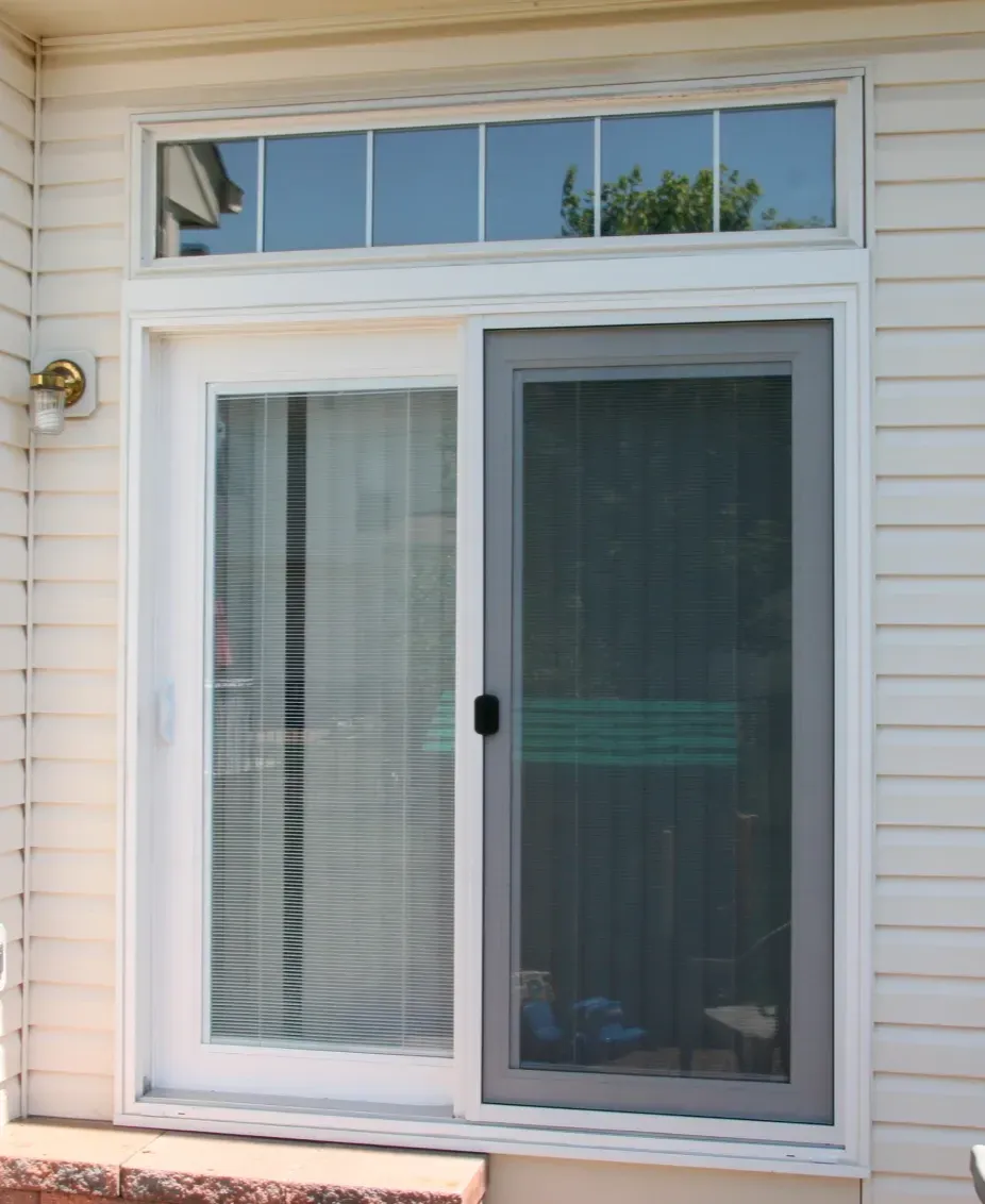 Sliding glass door with vertical blinds partially closed, set within white siding. A small rectangular transom window is above, and a light fixture is on the left.