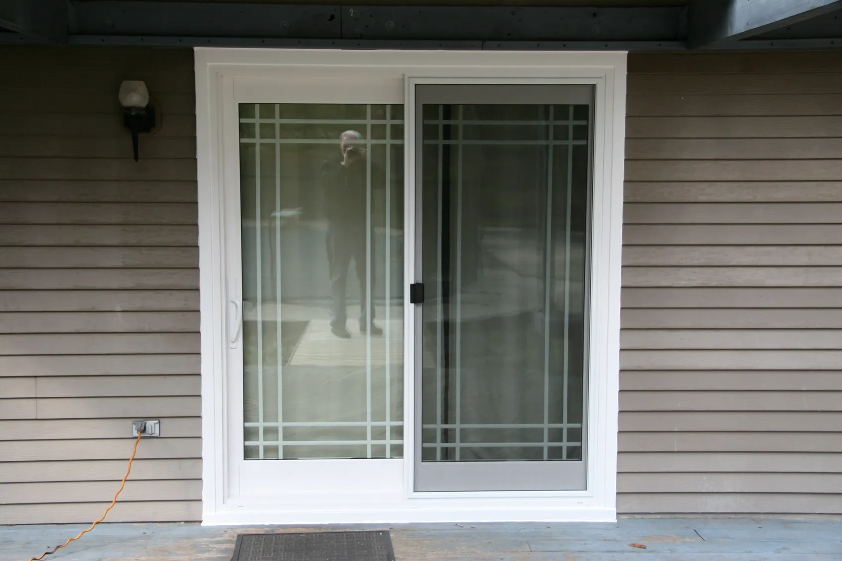 A glass sliding door with white gridded frame is set in a beige paneled wall. Reflection shows a person. A lamp and an orange cable are nearby.