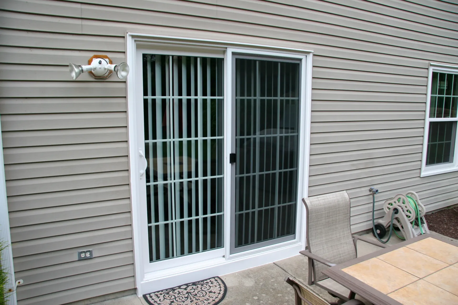 Sliding glass door with vertical blinds on a beige-sided house. Nearby, a patio table with chairs, wall light, window, and garden hose.