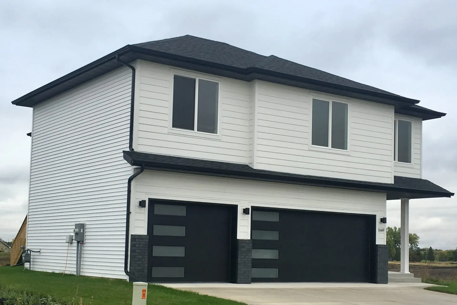 Modern two-story white house with black trim, featuring large windows and two black garage doors. Overcast sky and green lawn add a serene tone.