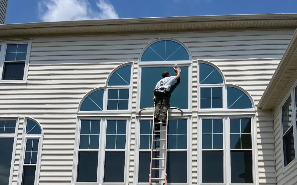 A person is on a ladder cleaning a large, multi-pane window with an arched design on a beige house under a clear blue sky, conveying focus and diligence.