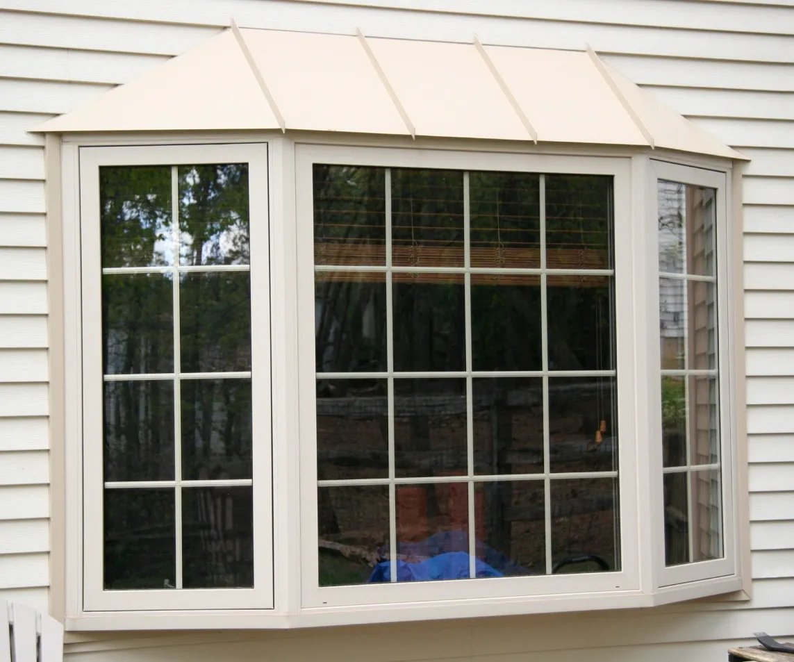 A beige bay window with four glass panels featuring grids, set in a vinyl siding wall. The window reflects greenery from outside, creating a serene ambiance.