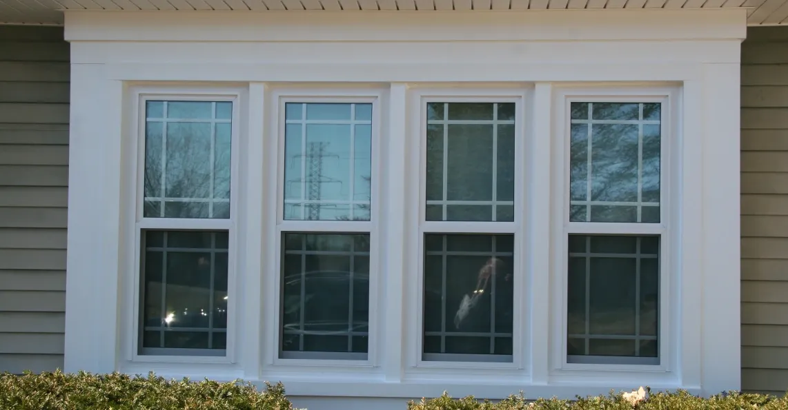 A set of three white-framed double-hung windows with grid patterns, reflecting daylight, is set against light gray siding. Bushes are below the windows.