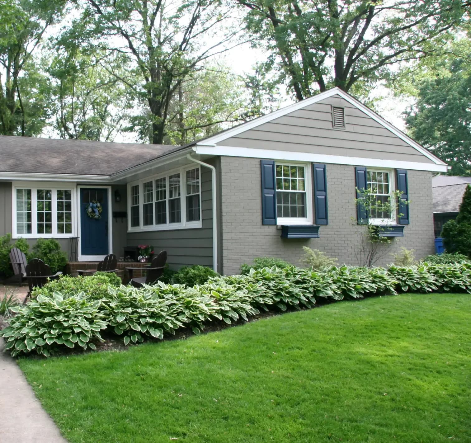 A two-story white colonial house with a red door, black shutters, large windows, and neatly trimmed shrubs on a lush green lawn. Overcast sky in the background.
