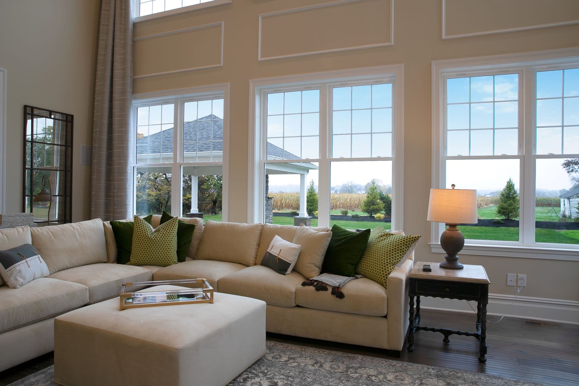 Spacious living room with large windows showing a garden view. Beige sectional sofa with green pillows, an ottoman, and a lit table lamp convey a cozy ambiance.